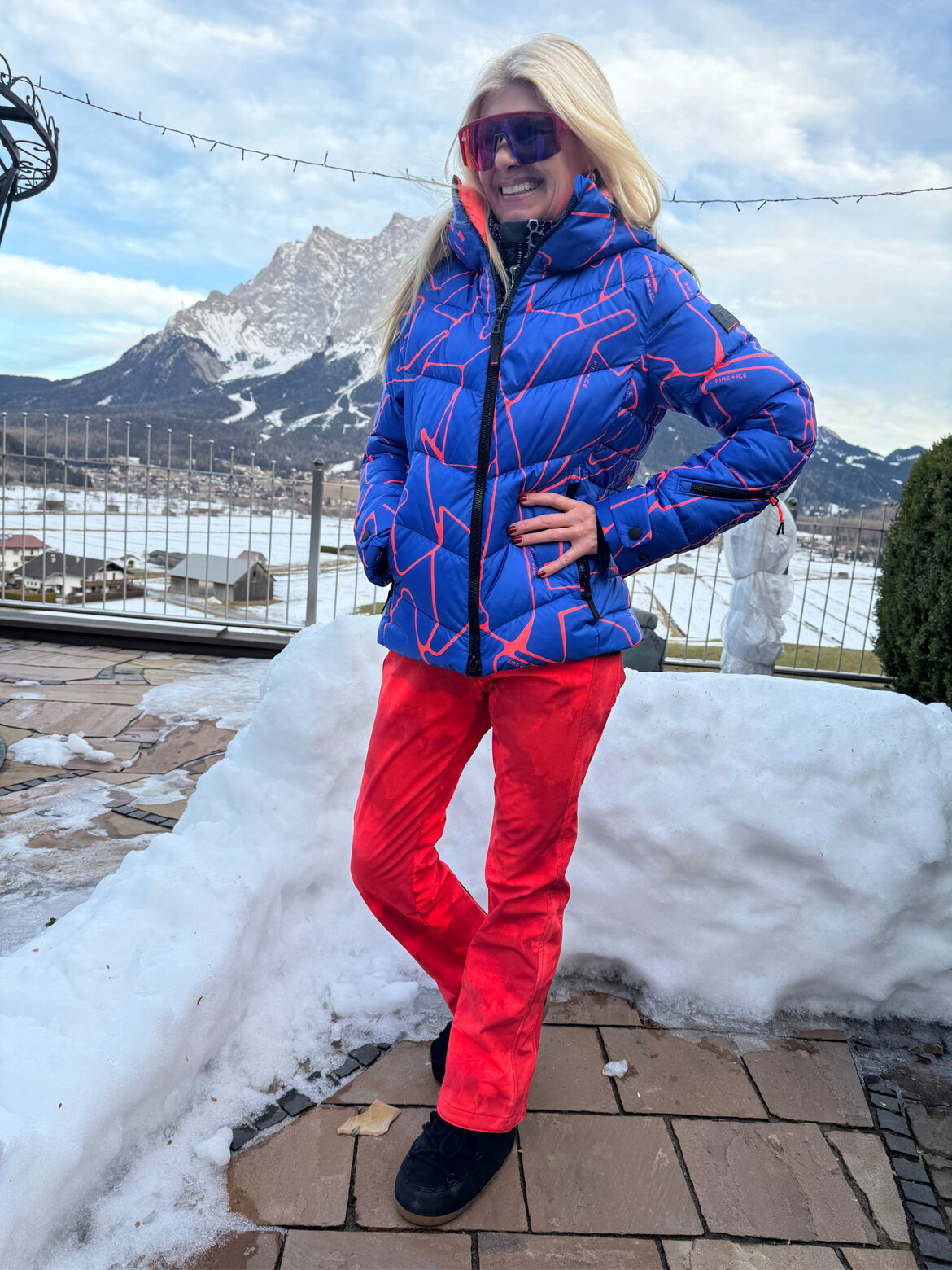 Renate Zott Ski-Outfits Knallfarben | © Renate Zott Renate Zott in blauer Daunenjacke mit roten Linien, roter Skihose und verspiegelter Brille posiert lächelnd vor verschneiter Bergkulisse. | © Renate Zott