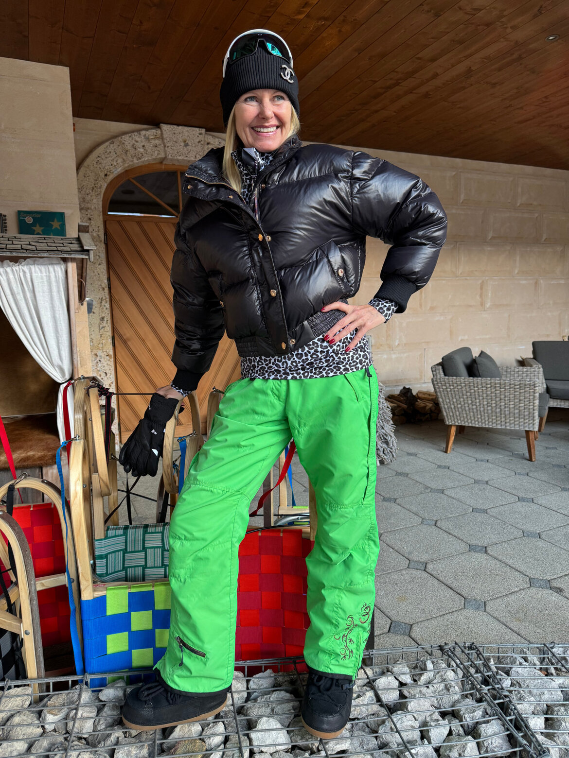 Renate Zott Ski-Outfits Snowboardhose | © Renate Zott Renate Zott in schwarzer Daunenjacke, grüner Skihose und Mütze mit Logo posiert lachend vor Holzschlitten mit bunten Sitzpolstern. | © Renate Zott