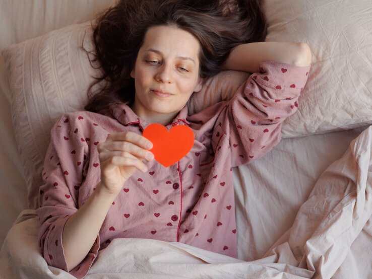 Eine Frau mit braunen Haaren liegt entspannt im Bett, trägt einen rosa Pyjama mit Herzmuster und hält ein rotes Papierherz in der Hand.