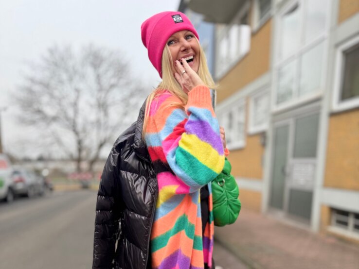 Renate Zott mit farbenfrohem Outfit, grüner Handtasche und schwarzen Plateau-Boots blickt strahlend über die Schulter.