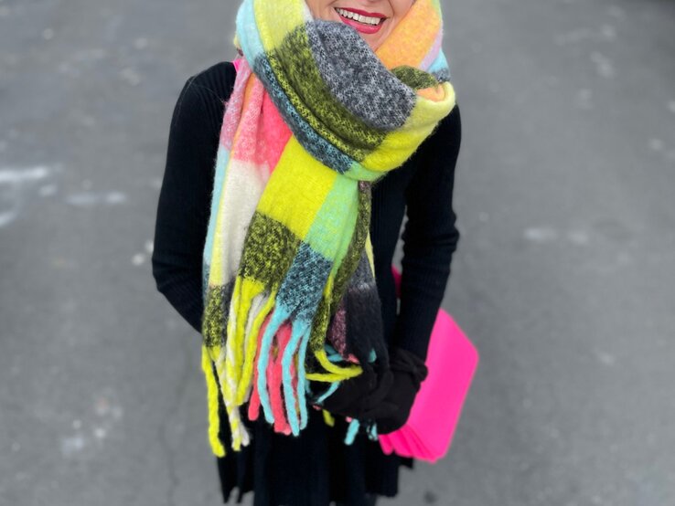 Renate Zott mit hochgestecktem Haar, buntem XXL-Schal und schwarzem Outfit steht auf einer Stadtstraße mit geparkten Autos.