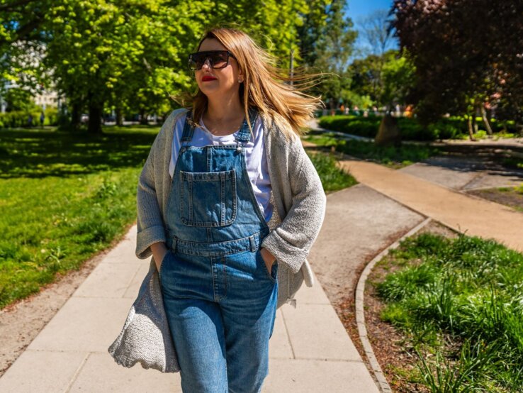 Frau in Latzhose und mit beiger Tasche genießt das Frühlingswetter in einem Park, umgeben von blühenden Bäumen und sattgrünem Gras.