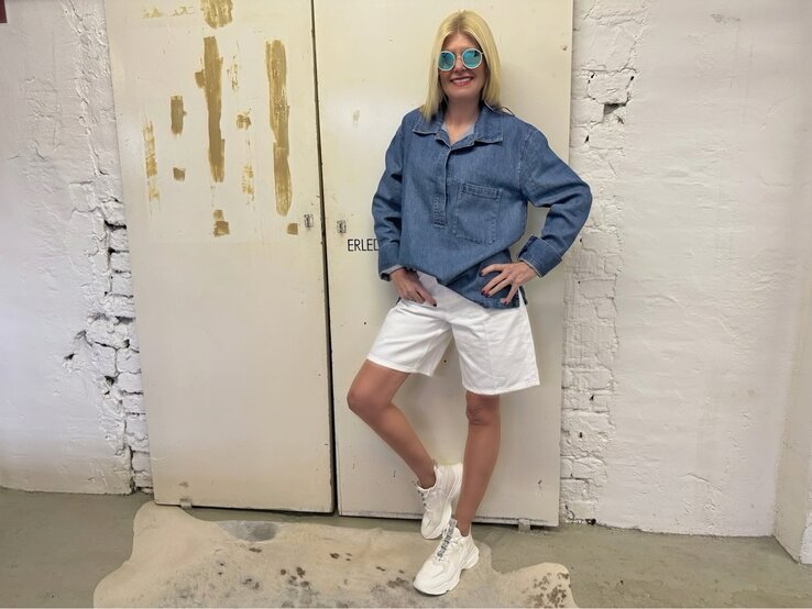 Mittelalte Frau mit glattem blondem Haar, Jeanstunika und runder Sonnenbrille posiert entspannt vor abgenutzter Tür.