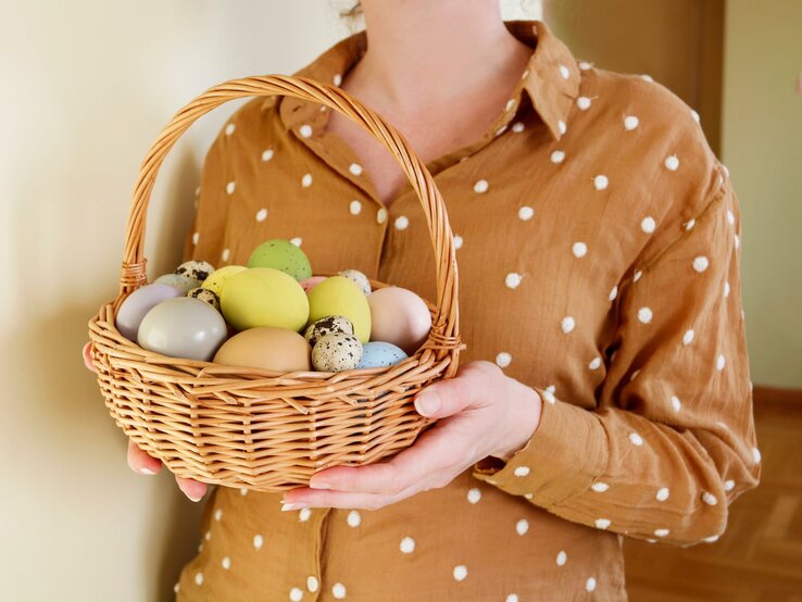 Ostern Brunch Bluse Tchibo Nahaufnahme einer Frau in gepunkteter Bluse, die einen geflochtenen Korb mit dekorierten Ostereiern präsentiert.