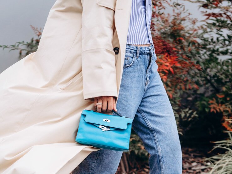 Zu sehen ist eine Frau, die einen Trenchcoat, einen kurzen dünnen Cardigan, eine Straight leg Jeans und eine hellblaue Tasche trägt.