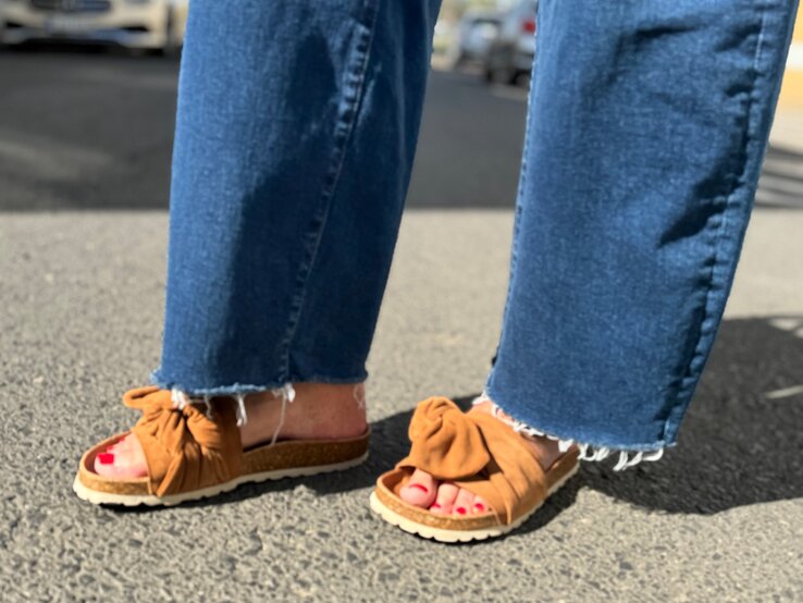 Füße einer Frau in braunen Sandalen mit Korksohle und rotem Nagellack auf Asphalt, Jeans mit ausgefranstem Saum.