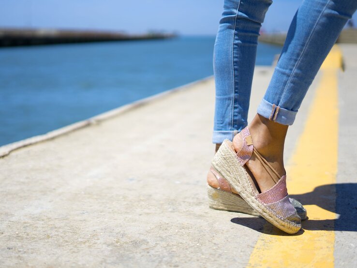 Beine einer Frau in hellblauen Jeans und rosafarbenen Espadrilles auf Betonweg neben blauem Wasser bei strahlendem Sommerwetter.