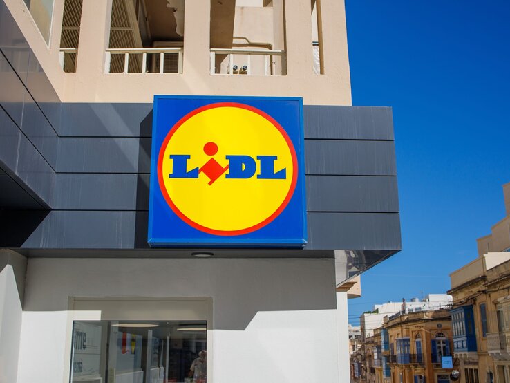 Lidl Schild im Sommer Leuchtendes Lidl-Schild mit gelbem Kreis auf blauem Hintergrund an moderner Fassade unter wolkenlosem Himmel.