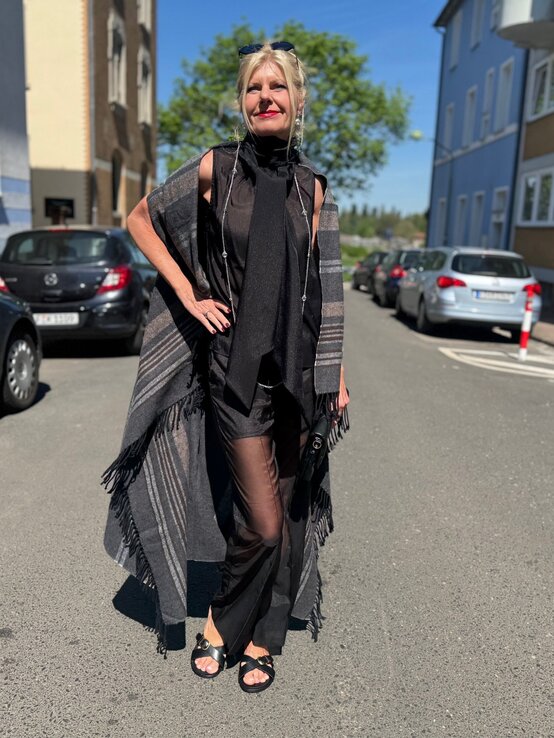 Renate Zott im komplett schwarzen Look mit Glanztop, transparenter Hose und grauem Fransencape unter blauem Himmel. | © Renate Zott