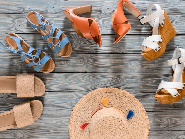 Verschiedene Damen-Sommersandalen in Beige, Blau, Orange und Weiß sowie ein Strohhut mit bunten Quasten liegen auf einem Holzboden.
