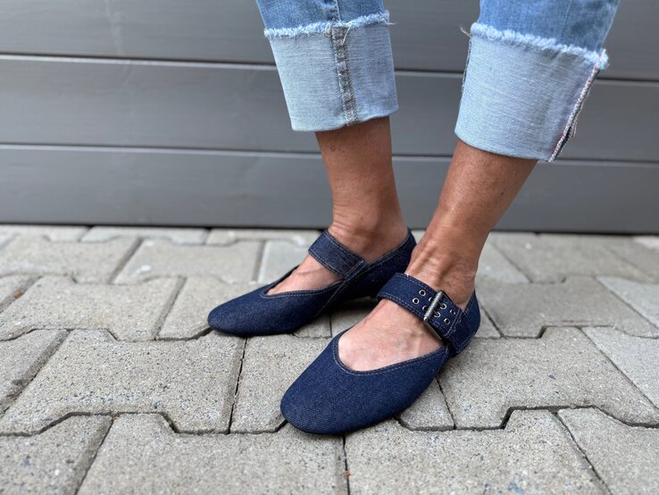 Frauenfüße in dunkelblauen Jeans-Ballerinas mit Schnalle auf grauem Pflaster, darüber ausgefranste, hochgekrempelte Jeans. | © Renate Zott