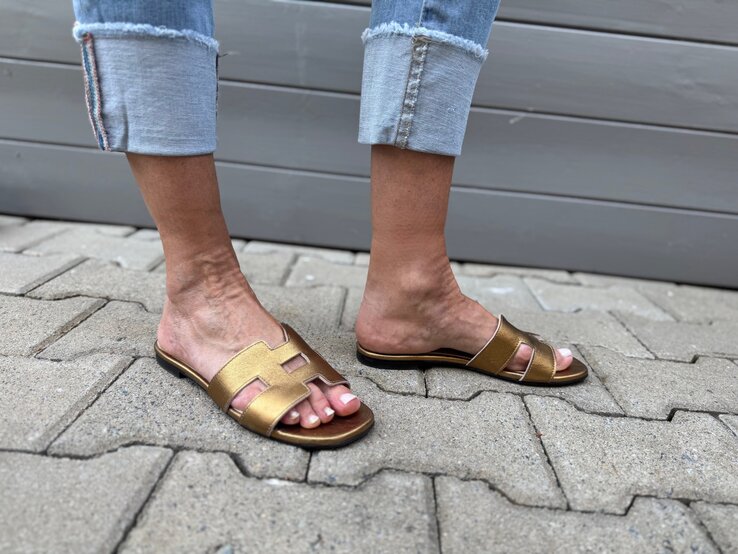 Gebräunte Füße in goldenen H-Sandalen auf grauem Steinpflaster, kombiniert mit hochgekrempelter Fransenjeans. | © Renate Zott