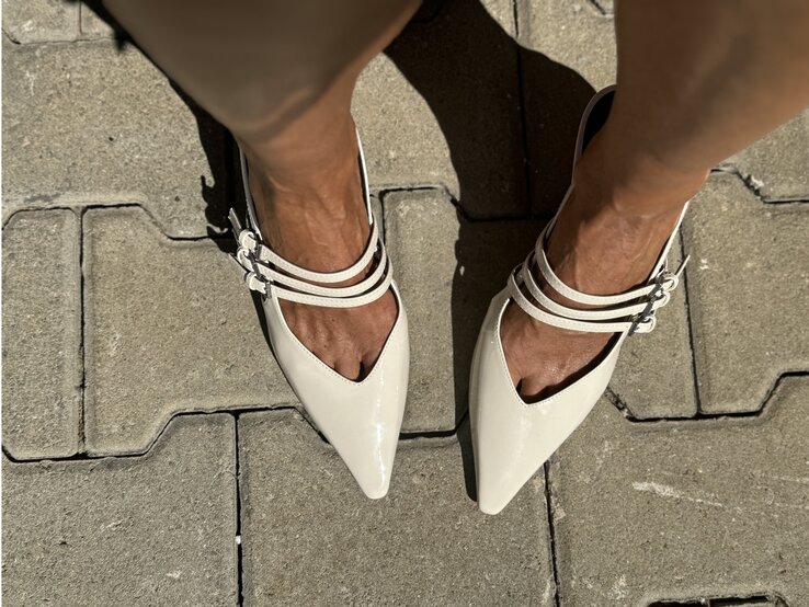 Füße in cremefarbenen Kitten Heels mit spitzer Kappe und drei Riemen auf sonnenbeschienenem Pflastersteinboden. | © Renate Zott