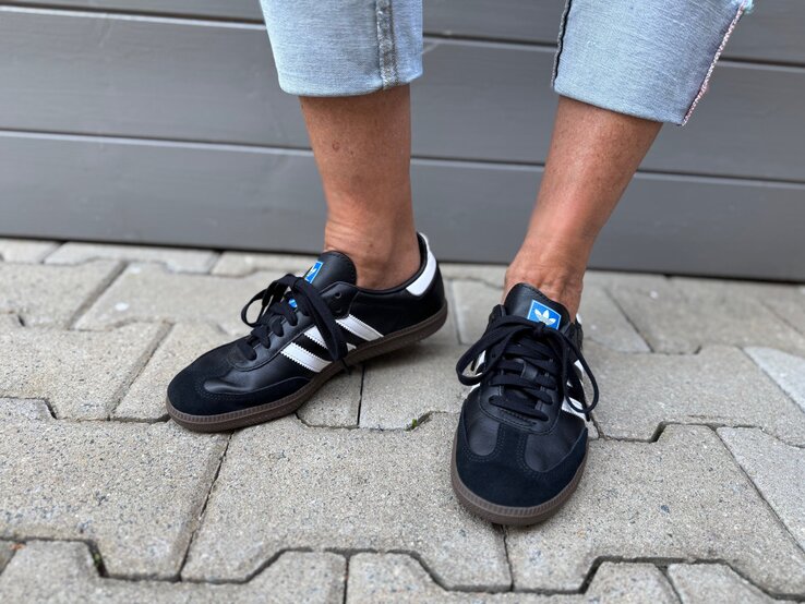 Schwarze Adidas-Sneaker mit weißen Streifen an gebräunten Füßen in gekrempelten Jeans auf grauem Pflasterstein. | © Renate Zott