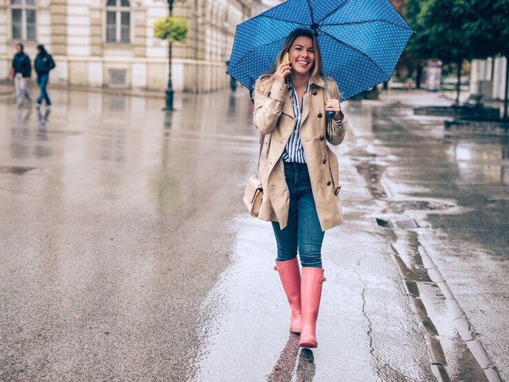 Lachende Passantin mit Regenschirm und Gummistiefeln spaziert durch regennasse Altstadtstraße mit glänzendem Pflaster.