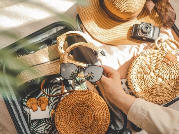 Offener Reisekoffer gefüllt mit Strandkleidung, Sommer-Sandalen, runder Basttasche, Sonnenhut und Hand mit schwarzer Sonnenbrille mit Pflanzenreflex.