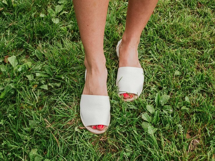 Weibliche Füße mit rotem Nagellack in weißen Espadrilles-Sandalen stehen auf frisch gemähtem, saftig grünem Rasen.