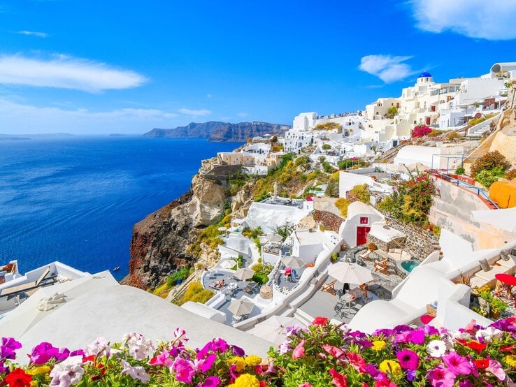Sandalen Luxus Griechenland Urlaub Griechische Küstenstadt mit strahlend weißen Häusern auf Klippen, Meerblick unter klarem Himmel, vorn blühende Petunien.