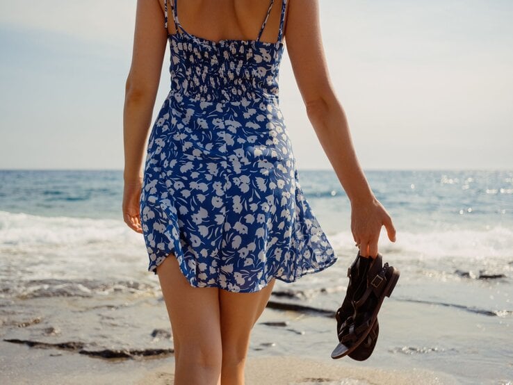 Sommerliche Szene am Meer: Frau im flatternden Kleid spaziert barfuß auf nassem Sand, Sandalen in der Hand.
