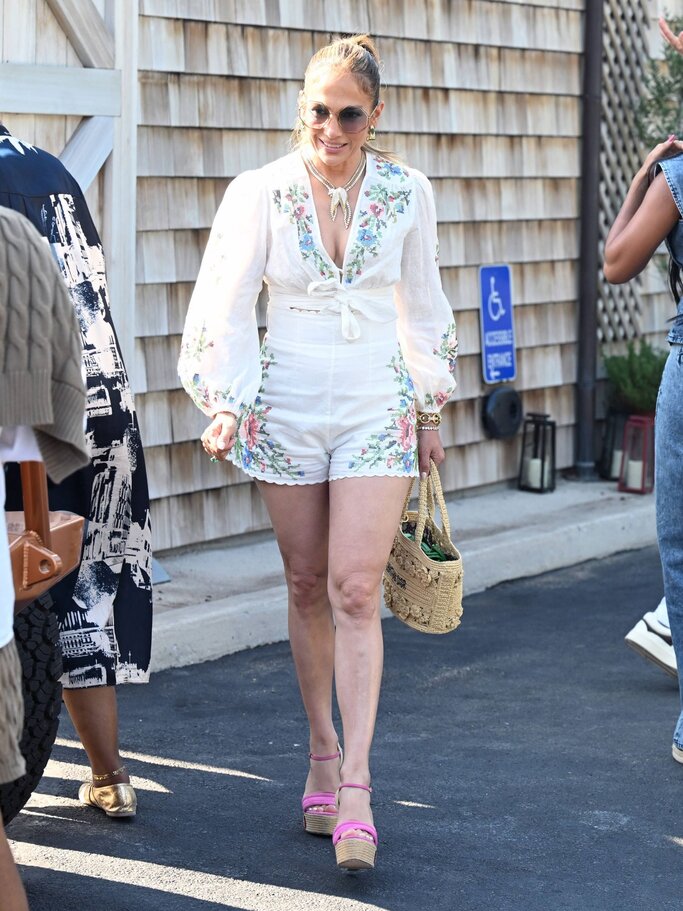 Jennifer Lopez mit strengem Pferdeschwanz trägt weißen bestickten Einteiler, rosa Keilsandaletten und Basttasche bei Sonnenschein. | © IMAGO / Cover-Images