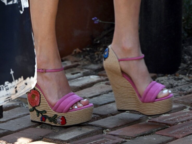 Detailaufnahme pinker Wedges mit Bastsohle und bunter Blumenstickerei auf rotem Pflastersteinboden. | © IMAGO / Cover-Images