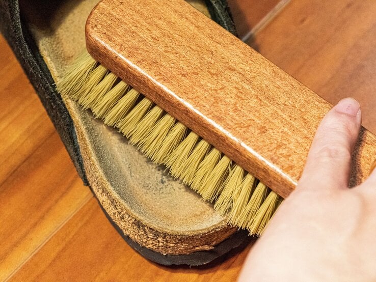 Hand reinigt mit einer Holzbürste die Innensohle einer getragenen Sandale mit Korkfußbett auf einem Holzfußboden.