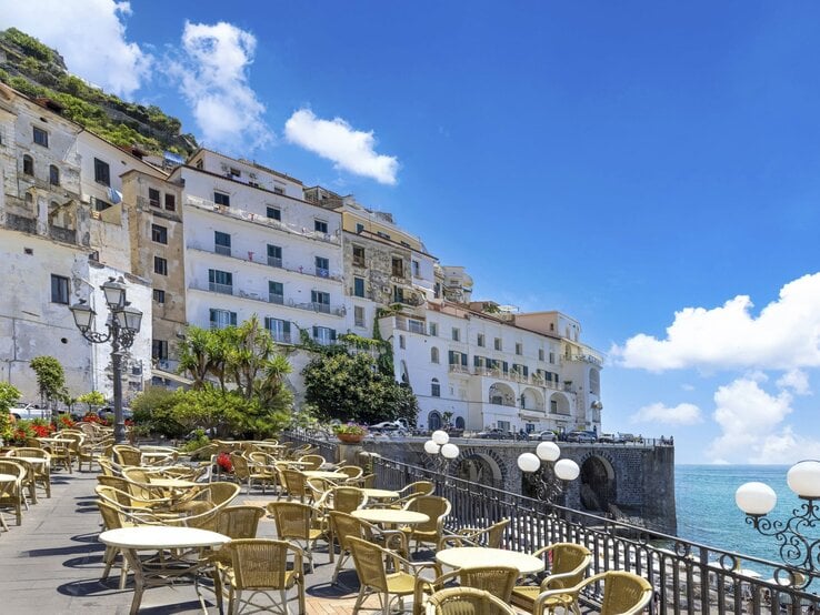 Sonnenbeschienene Terrasse mit leeren Rattanstühlen und Blick auf weiße Küstenhäuser und das tiefblaue Meer an der Amalfiküste.