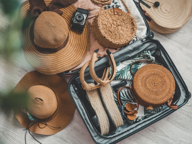 Flache Basttaschen, große Sonnenhüte und ein Koffer mit Palmenmuster-Badeanzug und Sandalen signalisieren sommerliche Urlaubsplanung.