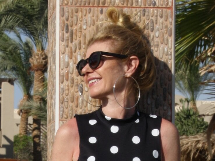 Anti-Aging-Accessoires: Renate Zott  lehnt an Mosaiksäule, trägt schwarze Sonnenbrille, große silberne Kreolen und ein gepunktetes Sommerkleid. | © Renate Zott