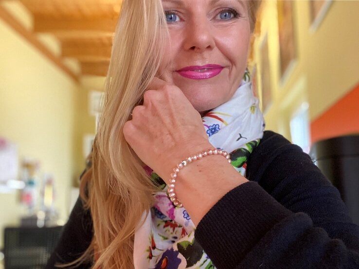 Anti-Aging-Accessoires: Renate Zott mit pinkem Lippenstift lächelt leicht und zeigt ein zartes Perlenarmband an ihrem Handgelenk. | © Renate Zott