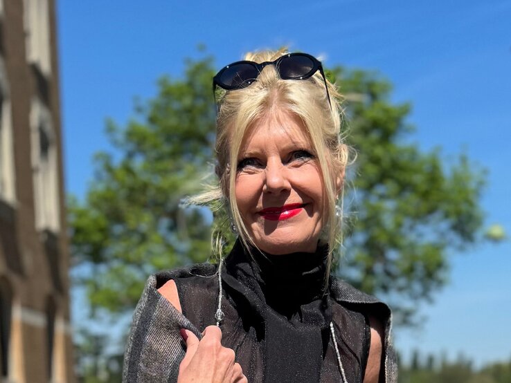 Anti-Aging-Accessoires: Renate Zott mit Hochsteckfrisur, rotem Lippenstift und schwarzer Sonnenbrille im Haar steht lächelnd vor blauem Himmel. | © Renate Zott