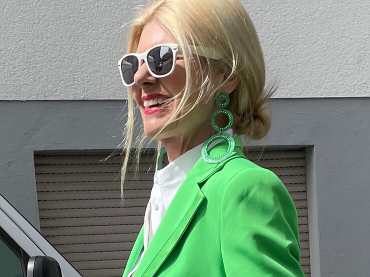 Anti-Aging-Accessoires: Renate Zott mit weißer Sonnenbrille und großen grünen Statement-Ohrringen trägt lächelnd einen knallgrünen Blazer. | © Renate Zott
