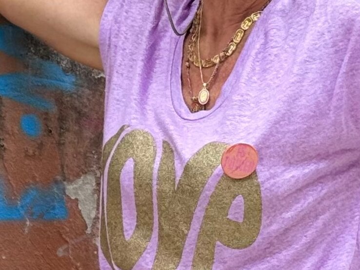 Anti-Aging-Accessoires: Detailansicht eines femininen Looks von Renate Zott mit goldenen Ketten und lila T-Shirt vor verwitterter Mauer mit blauer Graffiti. | © Renate Zott
