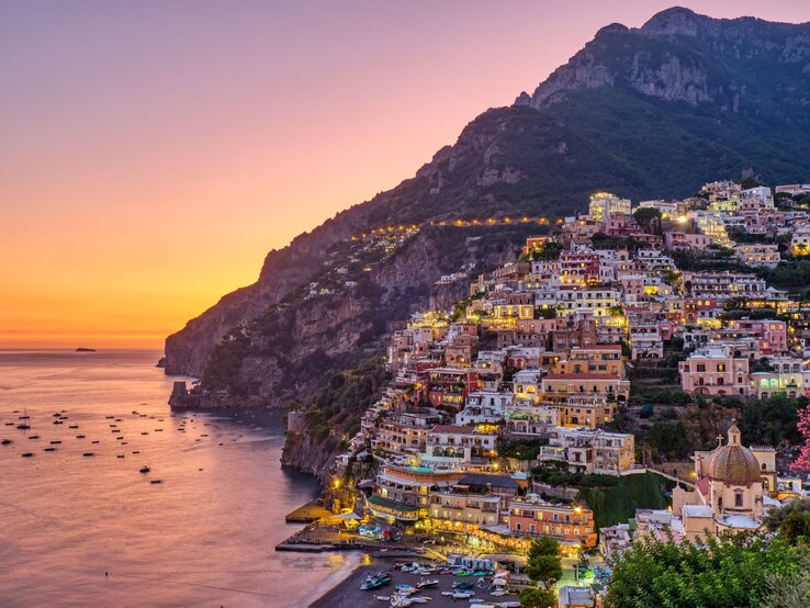 Symbolbild für Sandalen: Abendstimmung an der Amalfiküste. Beleuchtete Häuser von Positano schmiegen sich malerisch an steile Felsen über ruhigem Meer.