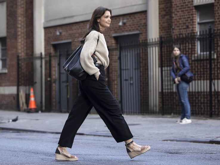 Katie Holmes in weitem Strickpullover, schwarzer Stoffhose und Espadrilles läuft lächelnd über eine Großstadtstraße.