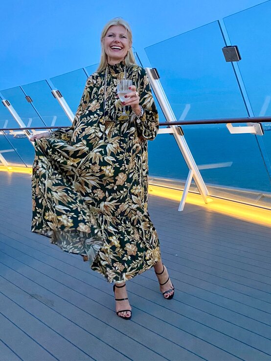 Elegant gekleidete Renate Zott mit Blumenmusterkleid und Riemchensandalen tanzt beschwingt auf einem Kreuzfahrtschiff bei Dämmerung. | © Renate Zott 
