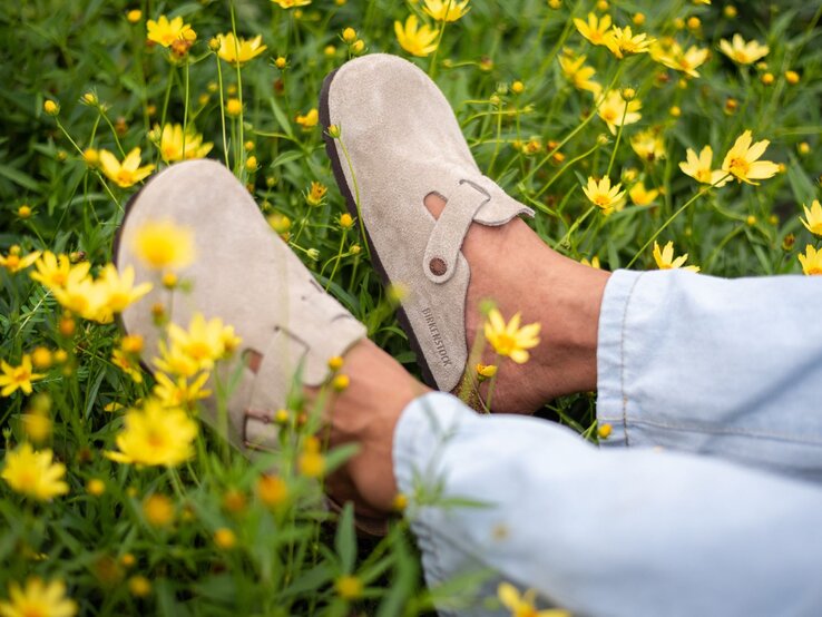 Bequeme Birkenstock-Clogs an braunen Füßen liegen locker in einem Feld aus sattgrünem Gras und leuchtenden Gelbblumen.