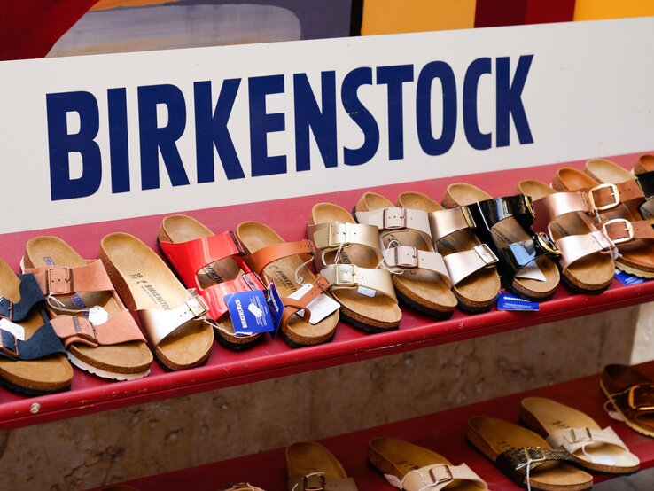 Bunte Birkenstock-Sandalen mit Korksohle stehen ordentlich nebeneinander auf einem roten Regal vor großem Firmenschild.