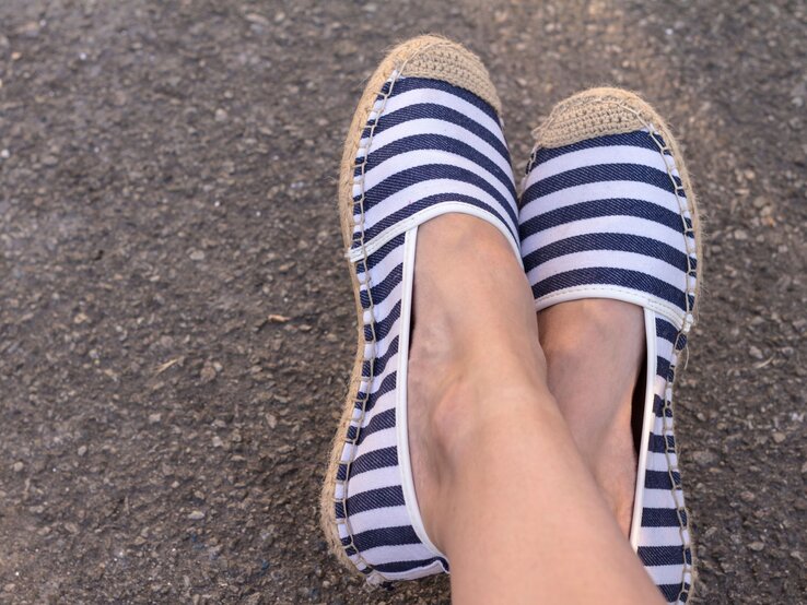 Blau-weiß gestreifte Espadrilles mit heller Kappe und Bastsohle an nackten Beinen auf grauem Straßenbelag.