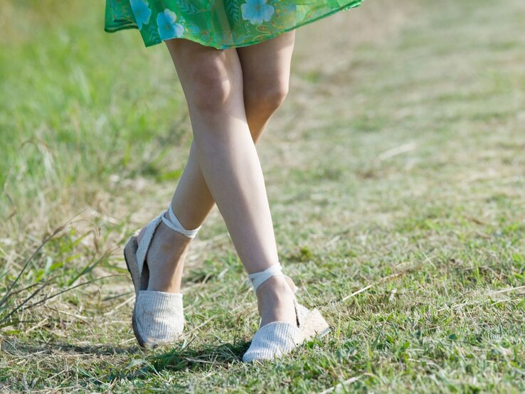 Junge Frau mit gebräunten Beinen geht barfuß in weißen Espadrilles-Sandalen über einen sonnigen Grasweg, grünes Sommerkleid weht leicht.