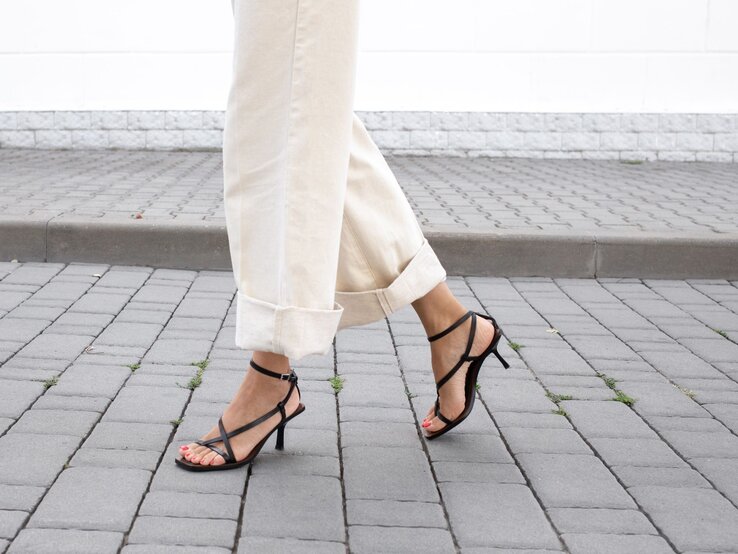 Sandalen Mode Trend Hose Eine Frau mit hochgekrempelter beigefarbener Hose geht auf Pflastersteinen in schwarzen Riemchensandalen mit Absatz.