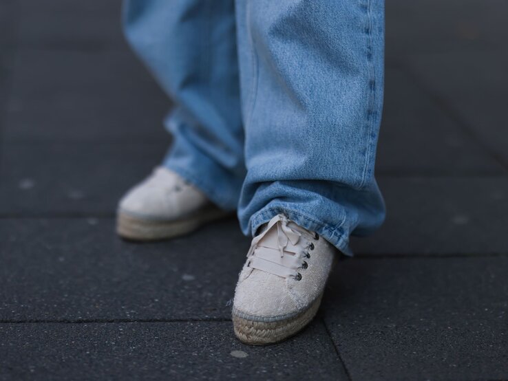 Espadrilles Sneaker Sommer Only Eine Person in weiten, bodenlangen Jeans trägt cremefarbene Espadrilles Sneaker auf dunkel gepflastertem Gehweg.