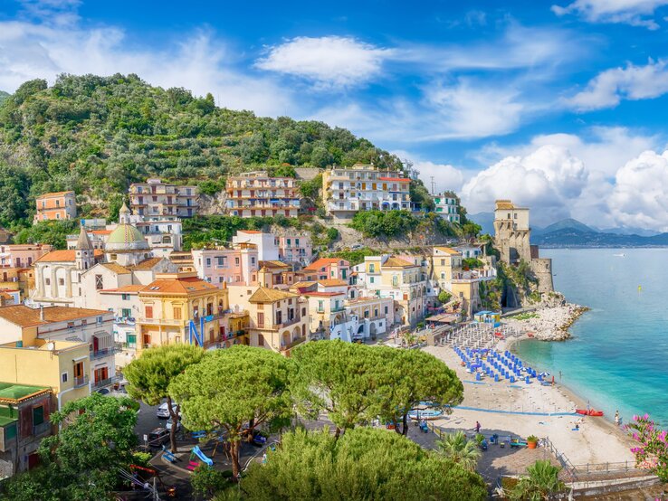 Amalfiküste Sandalen Sommer Urlaub Blick auf ein buntes Küstendorf mit grünen Hügeln, historischem Turm und einem ruhigen Strand unter leicht bewölktem Sommerhimmel an der Amalfiküste.