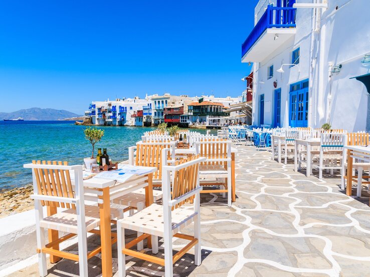 Weiß-blaue Taverne in Griechenland mit leeren Holztischen direkt am türkisfarbenen Meer, im Hintergrund bunte Balkone über dem Wasser.