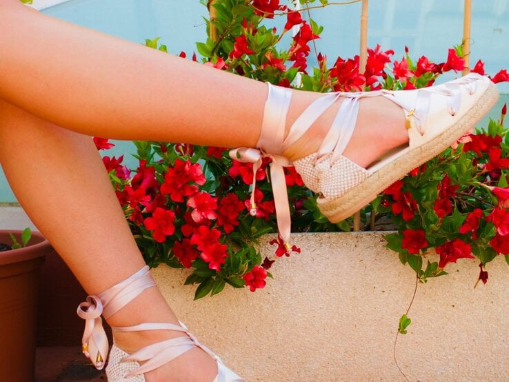 Zwei Beine in cremefarbenen Espadrilles mit glänzenden Satinbändern vor leuchtend roten Blüten in einem weißen Pflanzkübel.