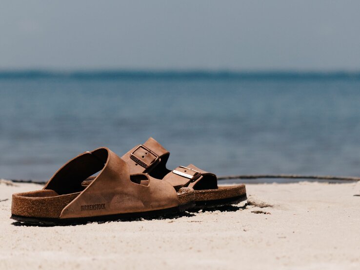 Ein Paar braune Birkenstock-Sandalen steht im hellen Sand vor ruhigem, türkisblauem Meer unter klarem Himmel.