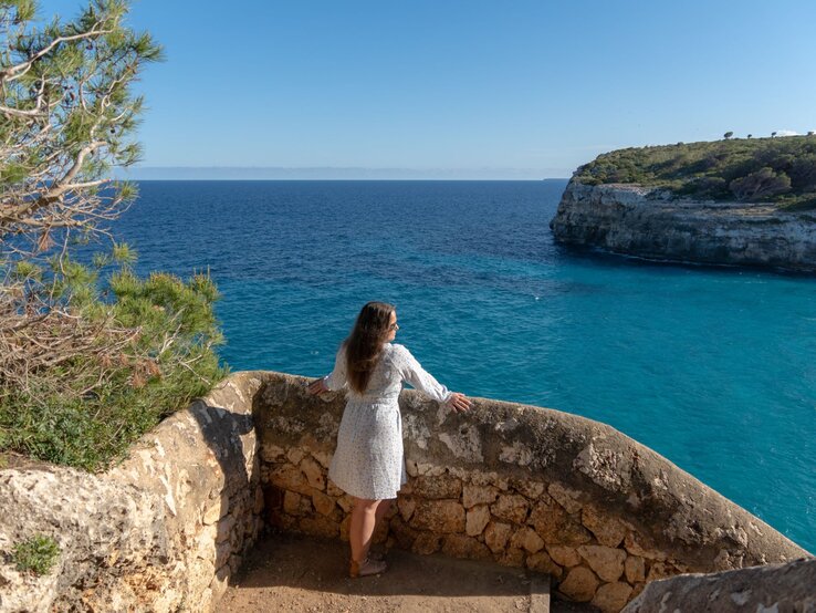 Frau in hellem Sommerkleid und Sandalen blickt entspannt von steinerner Aussichtsplattform auf türkisblaues Meer und steile Küste auf Mallorca.