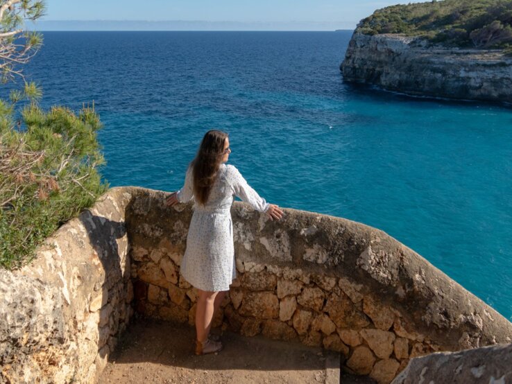 Frau in hellem Sommerkleid und Sandalen blickt entspannt von steinerner Aussichtsplattform auf türkisblaues Meer und steile Küste auf Mallorca.