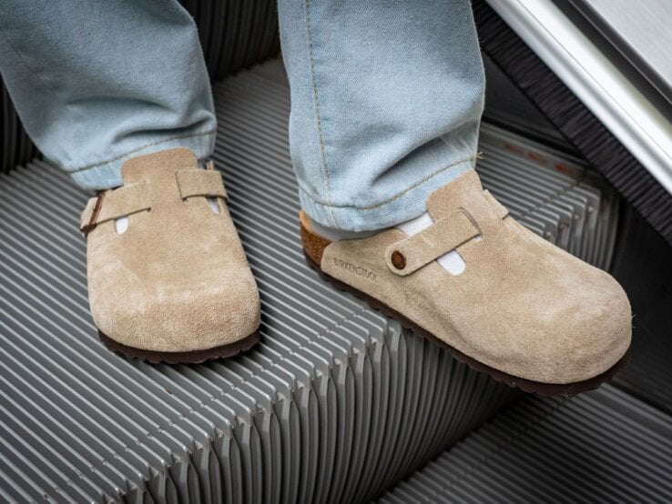Zwei Füße in beigen Wildleder-Clogs und weißen Socken stehen auf einer fahrenden Rolltreppe aus Metallrillen.