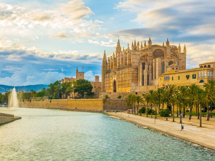 Die Kathedrale von Palma de Mallorca leuchtet im Abendlicht vor dramatischen Wolken, gesäumt von Palmen und einem ruhigen Wasserbecken.